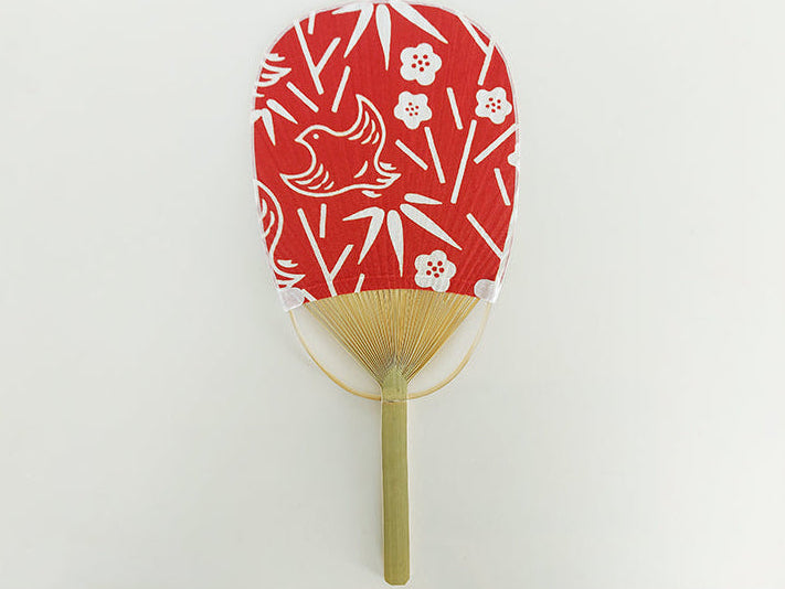 
                  
                    A traditional Japanese folding hand fan with a red paper panel decorated with white bamboo, crane, and plum blossom motifs, attached to a light bamboo handle.
                  
                